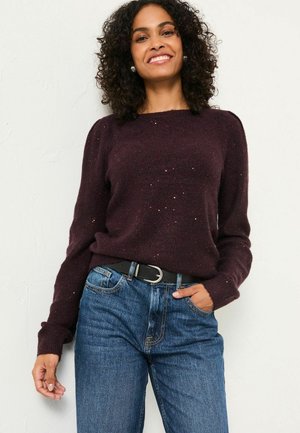 Frau mit lockigem Haar, die lächelt, trägt einen dunkelvioletten Pullover mit dezenten Glitzerakzenten, eine blaue Jeans und einen schwarzen Gürtel, und steht vor einer weißen Wand.