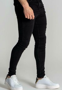 Svarta skinny jeans med en slät yta, framträdande sömmar och en åtsittande passform. Kombinerade med vita skor med gulddetaljer.