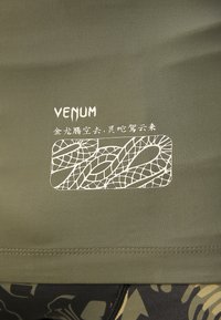 Grünes Sportoberteil mit gerippter Textur, das ein weißes Logo und ein abstraktes Designelement aufweist. Einfaches Design mit Nähten am Saum.
