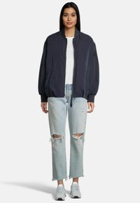 Marine bomberjack met een rits aan de voorkant, geribde manchetten en zakken, gecombineerd met lichtblauwe distressed jeans en grijze sneakers.