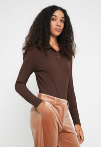 Mujer con cabello largo y rizado, vistiendo una camiseta marrón de manga larga con canalé y pantalones de terciopelo beige, de pie con una mano en el bolsillo.