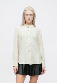 Camisa de botão de manga longa, na cor creme, com colarinho, apresentando botões em bronze, usada sobre uma saia pregueada preta.