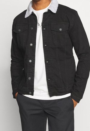 Mężczyzna ubrany w czarną dżinsową kurtkę z białym polarowym kołnierzem na białym T-shircie i ciemnych spodniach, z rękami w kieszeniach, wewnątrz.