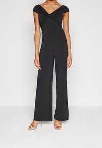 Schwarzer Jumpsuit mit einem verdrehten Vorderteil, kurzen Ärmeln und weitem Bein. Hergestellt aus einem glatten, elastischen Material; kombiniert mit Riemchensandalen.