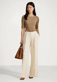 Nainen, jolla on päällään ruskea kuvioitu toppi, kermaiset leveälahkeiset housut, ruskea vyö, ruskeat kengät ja hän pitää ruskeaa käsilaukkua.