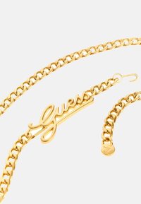 Gold-tone chain necklace featuring a cursive "Guess" logo charm. Thick, interlocking links with a hook closure and round branding tag.