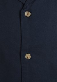 Mörkblå skjorta med texturerad väv, knäppning fram och en enda beige knapp med fyra hål på stången.