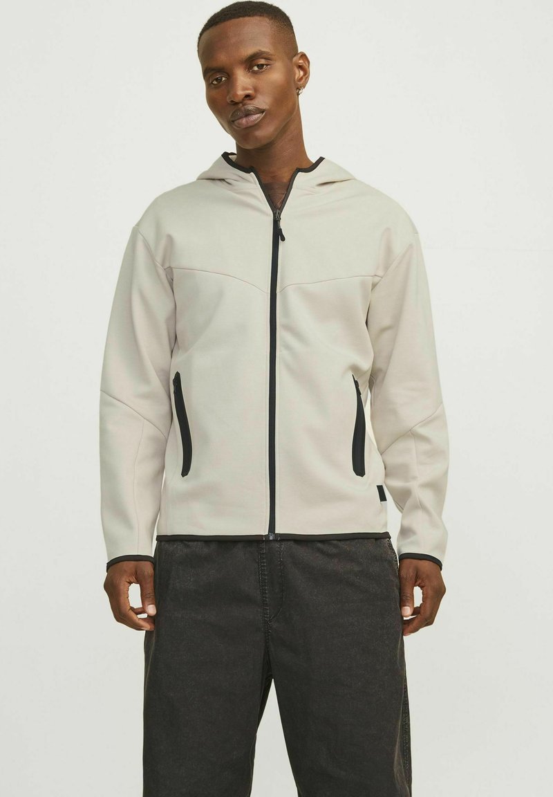 Veste zippée gris clair avec capuche, zips noirs et poches latérales ; présente une texture lisse et une coupe ajustée.