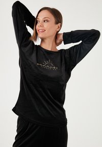 Zwart langemouwen top gemaakt van zachte stof, met een afgeronde zoom en een gouden zonlogo op de borst. Gladde textuur, relaxed fit.