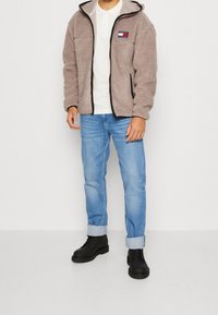 Chaqueta de felpa marrón con cremallera, detalles en negro y logo; camisa blanca debajo, jeans azules remangados y botas negras con cordones.