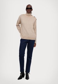 Beige kötött garbós pulóver bordás alsó résszel és gomb részlettel a vállon, sötétkék slim-fit farmernadrággal és fekete bokacsizmával párosítva.