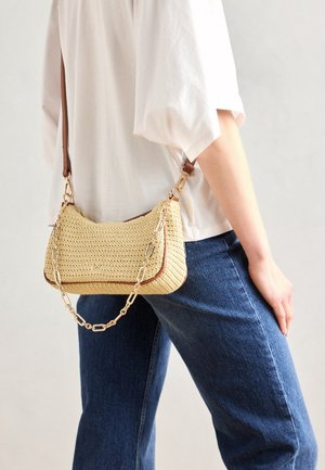 Bolso tejido amarillo con forma redondeada, ribetes marrones y correa de cadena. Exhibido sobre un fondo neutro junto a unos pantalones vaqueros.