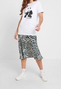 Vit bomull t-shirt med panda-grafik, tillsammans med en knälång, grön och krämfärgad djurmönstrad satin kjol, och vita sneakers.