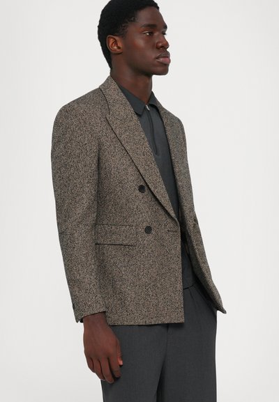 Man som bär en brun herringbone dubbelknäppt kavaj med svarta knappar över en mörkgrå zip-up skjorta och matchande byxor.