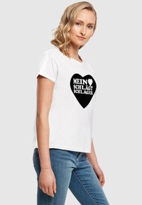 Hvid bomulds T-shirt med korte ærmer, med et sort hjerteformet design og hvid tekst der står "MEIN ♥ SCHLÄGT SCHLAGER."