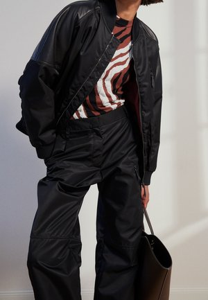 Persoon draagt een zwarte bomberjas, zwarte wijde broek, een bruine en witte zebraprint top, en houdt een grote zwarte handtas vast.