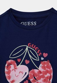 T-shirt in cotone navy con un design di cuore colorato con elementi rosa e rossi, logo "GUESS" e accenti a foglia. Collo rotondo.