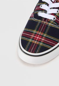 Sneaker din pânză în carouri, cu modele albastre, roșii și galbene, orificii albe și o talpă din cauciuc alb texturat cu margine neagră.