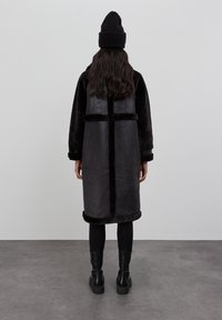 Schwarzer Kunstpelzmantel mit langer Silhouette, kontrastierender weicher Textur und umgeschlagenen Ärmeln. Getragen über schwarzen Leggings und klobigen schwarzen Stiefeln.