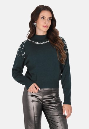 Donkergroene trui met een hoge hals en parelversieringen op de schouders. Gecombineerd met glanzende metallic zilveren broek.