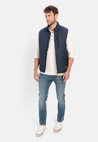 Marineblå quilted vest med høj krave, frontlynlås og sidelommer, parret med en hvid langærmet henley og blå jeans.