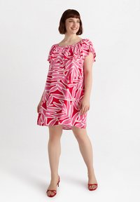Kurzes pinkes Kleid mit rötlich-pinken Blättern, rüschigem Ausschnitt und kurzen Ärmeln. Das Model steht in roten High Heels vor einem neutralen Hintergrund.