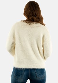 Maglione crema, morbido e soffice, con una vestibilità aderente e scollatura tonda, con decorazioni lungo la schiena. Abbinato a jeans blu scuro.