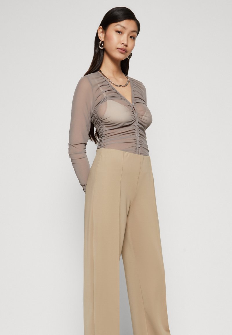 Durchsichtiger beiger Body mit Raffung und Knopfverschluss, kombiniert mit hoch geschnittenen beigen Hosen, die eine glatte Textur aufweisen.
