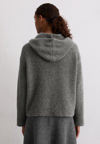 Szary, dziany hoodie z miękkiego, teksturowanego materiału. Posiada dużą kieszeń i długie rękawy, sięgające do talii. Subtelny, minimalistyczny design.