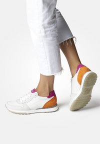Sneaker bianco con accenti in suede rosa e arancione, tomaia in pelle bianca texturizzata, suola piatta e design con lacci. Jeans bianchi a cropped visibili.