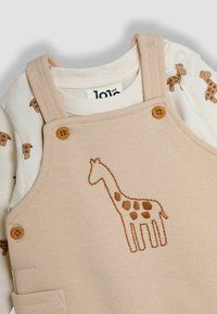 Beige tuinbroeken van textuurstof, met een geborduurde giraffe. Daaronder een witte shirt met lange mouwen met kleine bruine giraffe patronen.