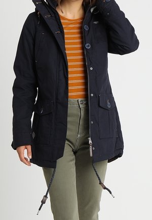 Personne portant une veste noire à capuche sur une chemise rayée orange et un pantalon vert clair, ajustant le col de la veste d'une main.