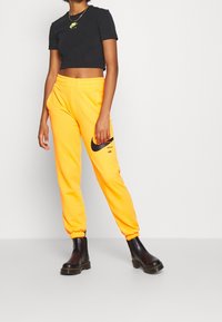 Černé zkrácené tričko s logem Nike, doplněné jasně žlutými tepláky s bočními kapsami a velkým černým logem Nike. Černé kotníkové boty.