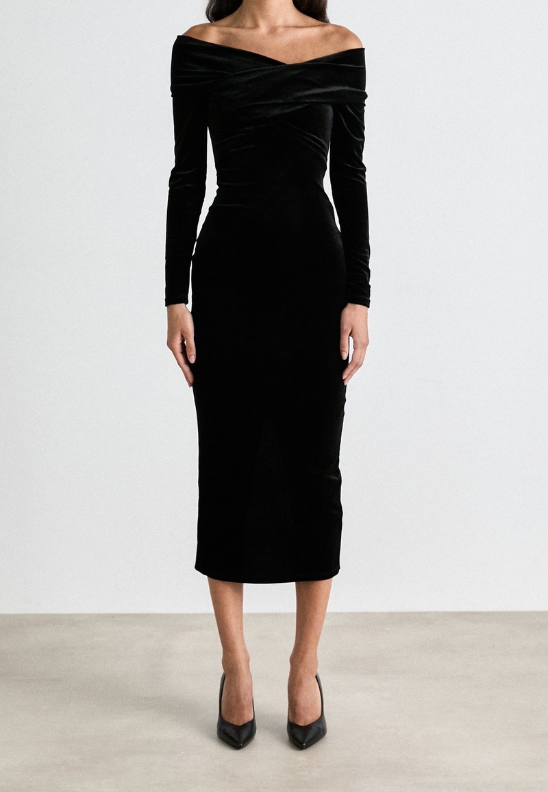 Femme portant une robe midi noire en velours moulante avec épaules dénudées, manches longues et talons noirs pointus, debout sur un sol gris.