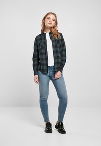 Flanellhemd im schwarz-türkisfarbenen Karomuster, mit Knopfleiste und zwei Brusttaschen. Kombiniert mit hellblauen engen Jeans und schwarzen Schuhen.