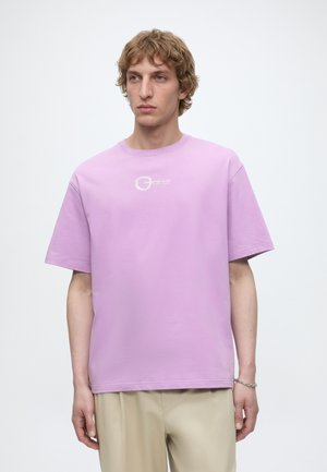 Jonge man met golvend haar die een lichtpaars T-shirt met kleine witte tekst en beige broek draagt, staand tegen een effen witte achtergrond.