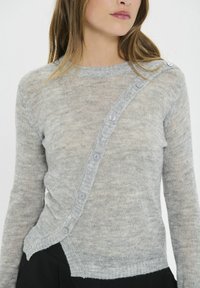 Maglione grigio chiaro lavorato a maglia con una texture leggermente trasparente, caratterizzato da una fila diagonale di bottoni e un orlo a coste.