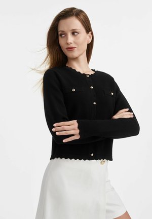 Czarny dziany kardigan z falbaniastymi krawędziami, złotymi guzikami i dwiema kieszeniami na piersi. Gładka tekstura, krótki krój i dopasowane rękawy.