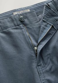 Pantaloni din bumbac ușor, de culoare albastră, cu fermoar la front, închidere cu nasture și detalii cusute, având eticheta de brand vizibilă pe talie.