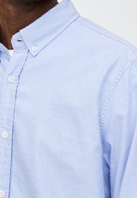 Hellblaues Hemd mit Knöpfen und Kragen. Hergestellt aus einem weichen Stoff mit glatter Textur und sichtbaren Nahtdetails.