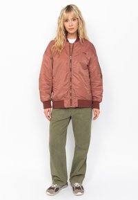 Chaqueta bomber marrón con un acabado suave y brillante, cierre de cremallera, puños y dobladillo de canalé. Pantalones holgados verdes oliva y zapatillas grises.