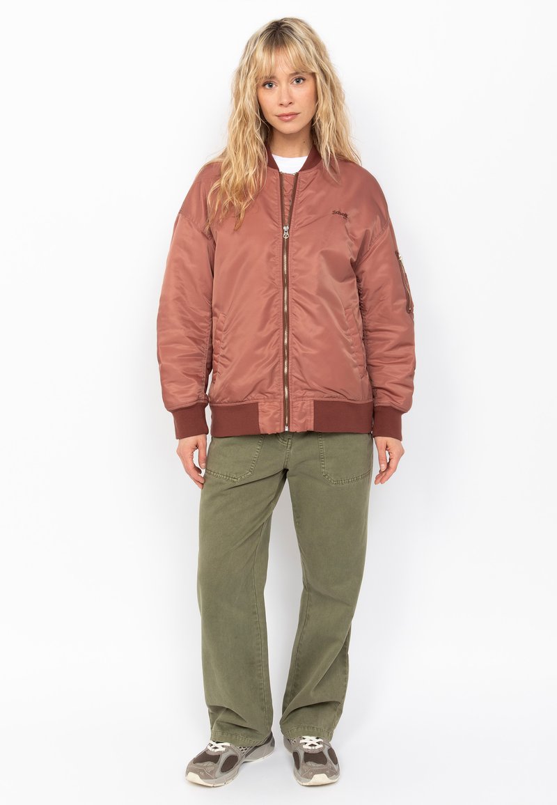Chaqueta bomber marrón con un acabado suave y brillante, cierre de cremallera, puños y dobladillo de canalé. Pantalones holgados verdes oliva y zapatillas grises.