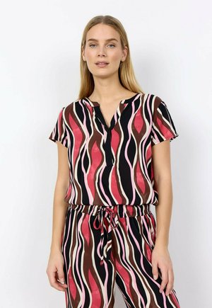 Vrouw die een bijpassende kortemouwen top en broek draagt met golvende strepen in rood, roze, bruin en zwart, staand tegen een witte achtergrond.