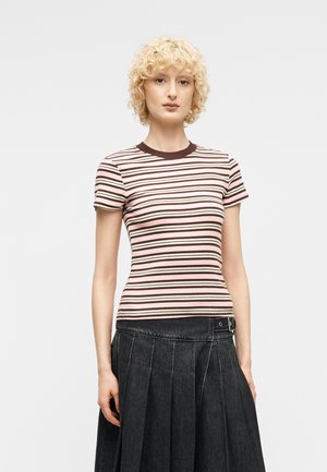 Νεαρό άτομο με κοντά ξανθά σγουρά μαλλιά που φοράει ροζ και καφέ ριγέ κοντομάνικη μπλούζα και σκούρο πλισέ φούστα με ζώνη με πόρπη.