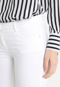 White jeans with a slim fit, featuring pockets, contrasting stitching, and a five-pocket design. Worn with a black-and-white striped blouse.