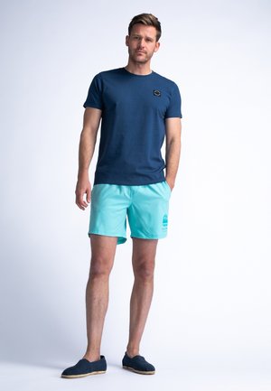 T-shirt di cotone blu navy con un piccolo logo, abbinato a pantaloni da bagno turchesi chiari con un disegno stampato. Espadrillas nere completano il look.