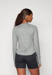 Top desportivo de manga comprida, cinza mesclado, com decote redondo, tecido texturizado e detalhes reflexivos discretos nas costas. Combinado com calções pretos.