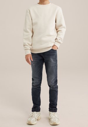 Personne portant un sweat-shirt beige uni, un jean bleu foncé légèrement usé et des baskets blanches, debout avec une main dans la poche.