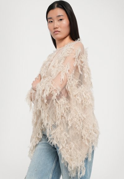 Beige, transparenter Poncho mit strukturierten Fransen, offener Front und weichem, fließendem Stoff, kombiniert mit hellblauen, verzierten Jeans.