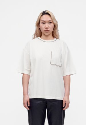 Giovane con lunghi capelli biondi che indossa una maglietta bianca oversize con decorazioni in strass sul colletto e sulla tasca, e pantaloni neri.